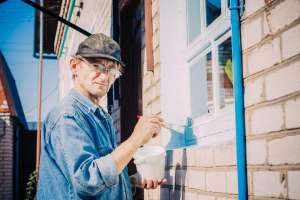 Caucasian Mature Man In Glasses And Cap Painting The Window Frame Outside Of The Private Brick House In Sunny Day.