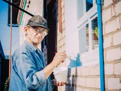 Caucasian Mature Man In Glasses And Cap Painting The Window Frame Outside Of The Private Brick House In Sunny Day.