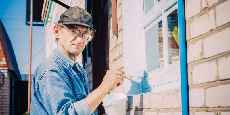 Caucasian Mature Man In Glasses And Cap Painting The Window Frame Outside Of The Private Brick House In Sunny Day.