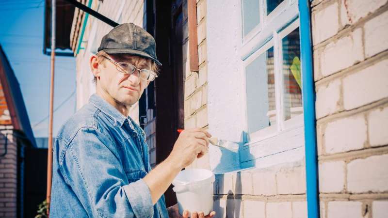 Caucasian Mature Man In Glasses And Cap Painting The Window Frame Outside Of The Private Brick House In Sunny Day.