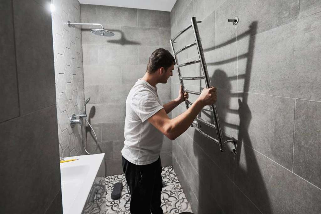 Male plumber hanging electric towel rack dryer on the wall in bathroom. Man installing stainless steel bath towel heater in apartment under renovation. Home renovation concept.