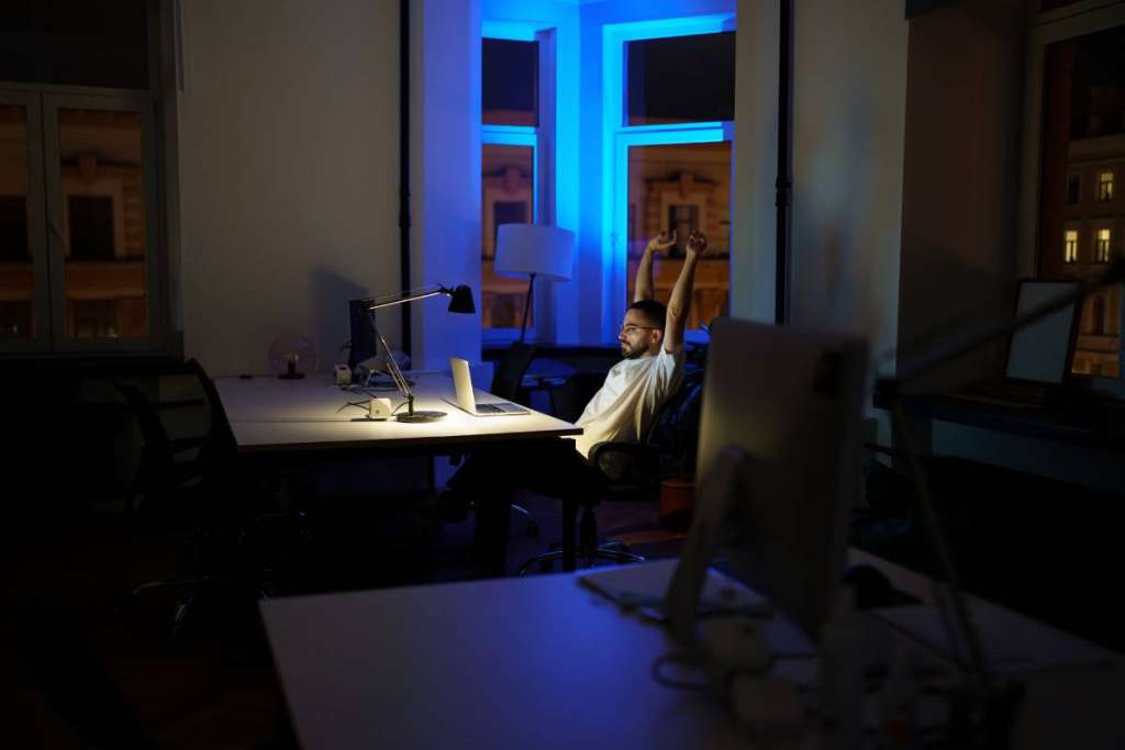 Male freelancer work late night stretching hands and back while sitting at laptop computer in dark office. Tired man programmer web designer overtime within deadline. Exhausted developer having break (Male freelancer work late night stretching hands a